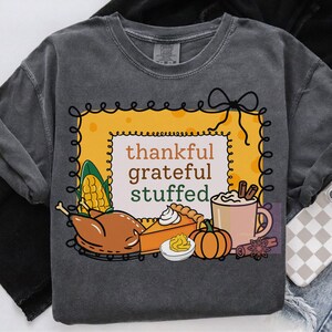 May include: Dark gray sweatshirt with a Thanksgiving-themed graphic. The design features a yellow frame with the words "thankful grateful stuffed." Illustrations include a turkey, pumpkin pie, pumpkin, corn, and a mug of hot cocoa.