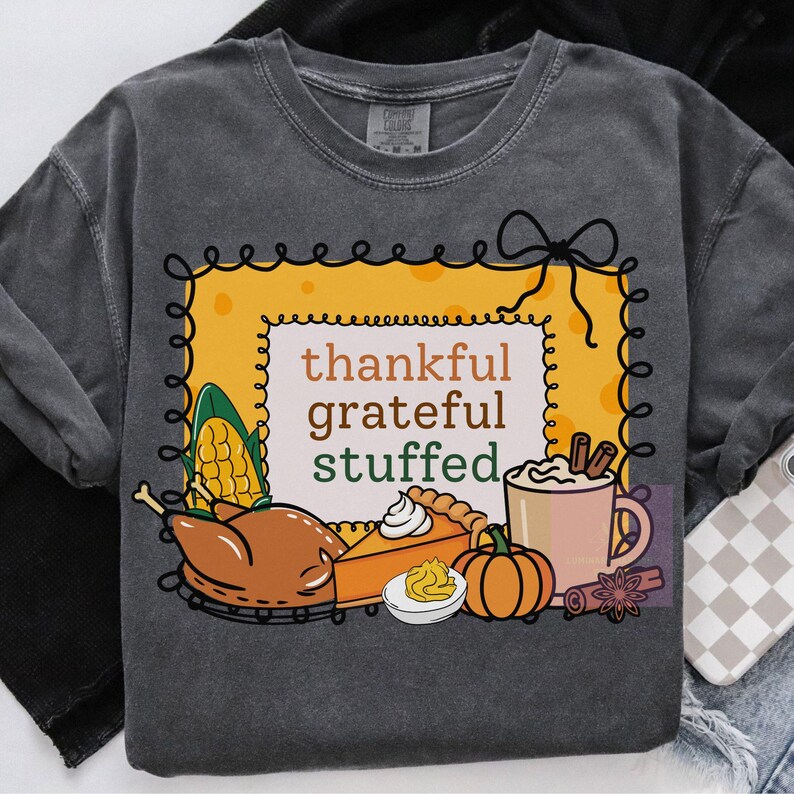 May include: Dark gray sweatshirt with a Thanksgiving-themed graphic. The design features a yellow frame with the words "thankful grateful stuffed." Illustrations include a turkey, pumpkin pie, pumpkin, corn, and a mug of hot cocoa.