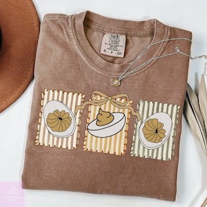 Op de afbeelding: Bruin Comfort Colors T-shirt met een afbeelding van drie gevulde eieren. Elk ei staat in een rechthoekig kader met verticale strepen. Een zilveren ketting met een kleine hanger ligt op het shirt.