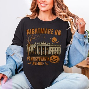 K&ouml;nnte beinhalten: Schwarzes Sweatshirt mit einem Halloween-Motiv. Das Design zeigt den Text "NIGHTMARE ON PENNSYLVANIA AVENUE" &uuml;ber einer Illustration eines Geb&auml;udes, eines Baumes, eines K&uuml;rbisses, Flederm&auml;usen und einem Mond.