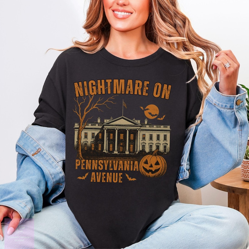 K&ouml;nnte beinhalten: Schwarzes Sweatshirt mit einem Halloween-Motiv. Das Design zeigt den Text "NIGHTMARE ON PENNSYLVANIA AVENUE" &uuml;ber einer Illustration eines Geb&auml;udes, eines Baumes, eines K&uuml;rbisses, Flederm&auml;usen und einem Mond.