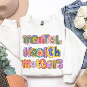 May include: A white sweatshirt with the words "Mental Health Matters" in colorful, bubble letters. The text is outlined in various colors, including pink, green, blue, and yellow. The sweatshirt is folded on a white surface.