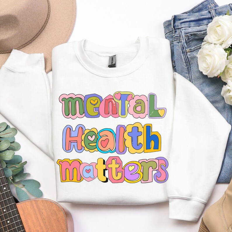 May include: A white sweatshirt with the words "Mental Health Matters" in colorful, bubble letters. The text is outlined in various colors, including pink, green, blue, and yellow. The sweatshirt is folded on a white surface.