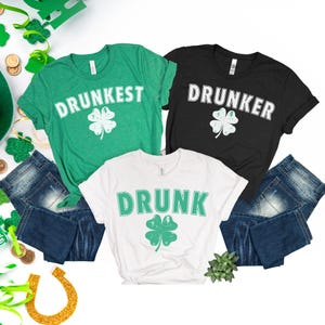 Może przedstawiać: Trzy koszulki z motywami Dnia Świętego Patryka. Zielona koszulka mówi "Drunkest", czarna koszulka mówi "Drunker", a biała koszulka mówi "Drunk". Każda koszulka ma koniczynę. Niebieskie dżinsy są złożone pod koszulkami.