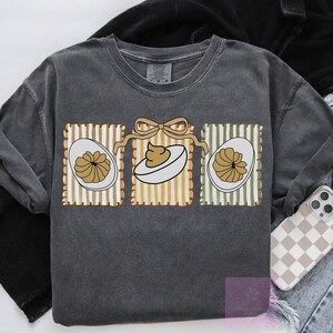 May include: Dark gray sweatshirt featuring a graphic of three deviled eggs. Each egg is in a rectangular frame with vertical stripes in brown, yellow, and green. A tan bow connects the frames. The sweatshirt is a Comfort Colors brand.