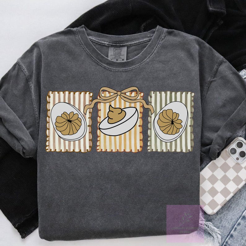 May include: Dark gray sweatshirt featuring a graphic of three deviled eggs. Each egg is in a rectangular frame with vertical stripes in brown, yellow, and green. A tan bow connects the frames. The sweatshirt is a Comfort Colors brand.
