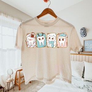 May include: A cream-colored t-shirt featuring a whimsical design of four cartoon teeth, each adorned with festive winter accessories. The teeth are framed in colorful rectangular boxes, with a Santa hat, reindeer antlers, a scarf, and a winter hat. The shirt is hanging on a wooden hanger.