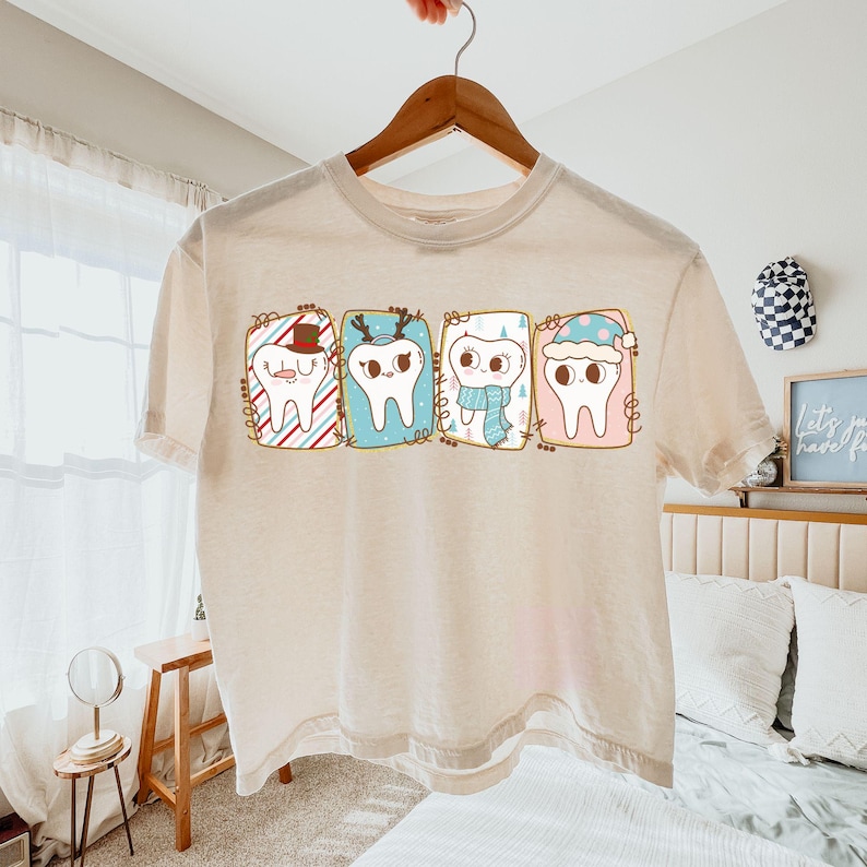 May include: A cream-colored t-shirt featuring a whimsical design of four cartoon teeth, each adorned with festive winter accessories. The teeth are framed in colorful rectangular boxes, with a Santa hat, reindeer antlers, a scarf, and a winter hat. The shirt is hanging on a wooden hanger.