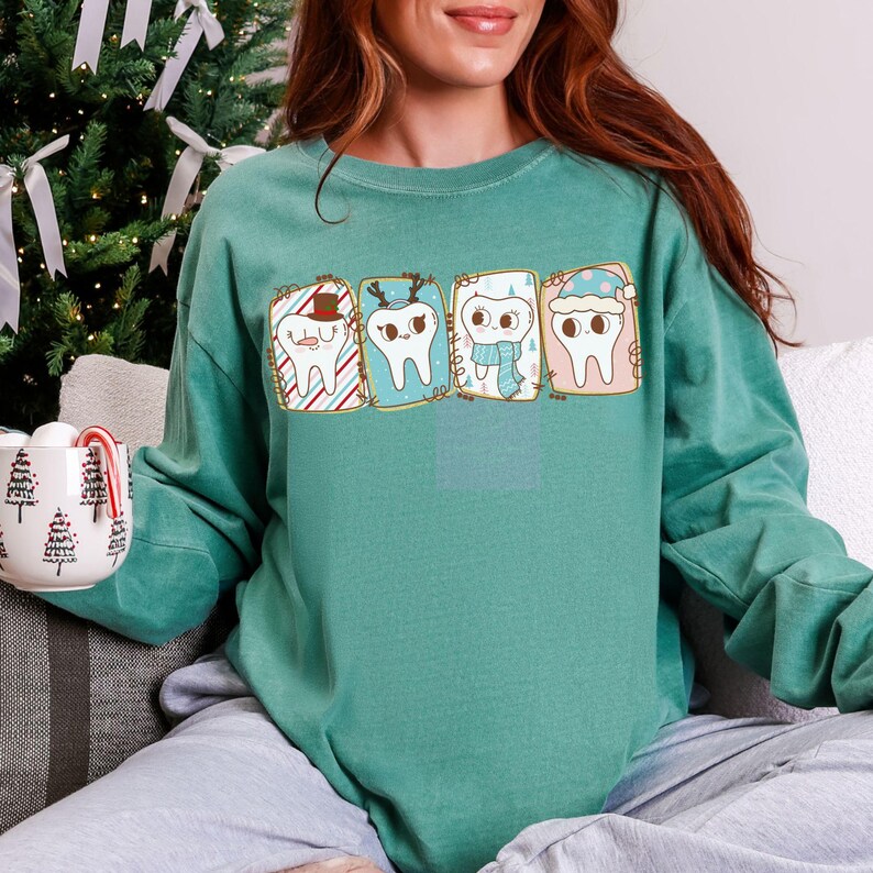 May include: Teal green sweatshirt featuring four cartoon teeth in festive holiday designs. Each tooth is inside a rectangular frame with a unique holiday theme, including a Santa hat, reindeer antlers, a scarf, and a winter hat.