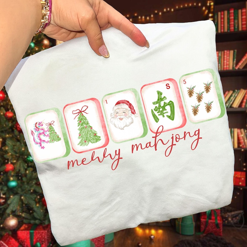 May include: A white sweatshirt featuring a graphic of five mahjong tiles with Christmas-themed illustrations including a dragon, a tree, Santa Claus, a green character, and pinecones, with the red cursive text merry mahjong below them.