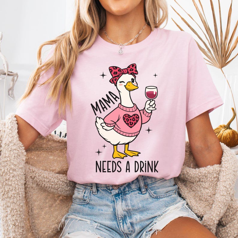 Peut inclure: T-shirt rose clair avec un dessin d'oie de dessin anim&eacute; portant un pull rose et un n&oelig;ud, tenant un verre de vin. Le texte "MAMA NEEDS A DRINK" est imprim&eacute; sur le t-shirt. L'oie a un motif de c&oelig;ur sur son pull.