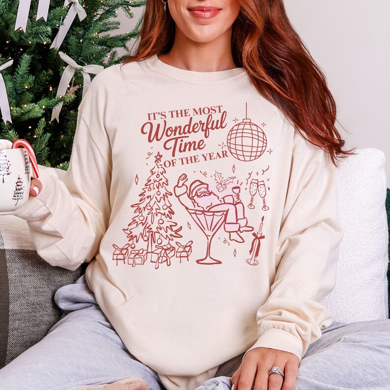 Puede incluir: Camiseta de manga larga color crema con un gr&aacute;fico festivo. El gr&aacute;fico presenta un &aacute;rbol de Navidad, Pap&aacute; Noel en una copa de Martini y el texto "It's the Most Wonderful Time of the Year" en borgo&ntilde;a.
