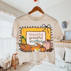 May include: Beige t-shirt with a Thanksgiving-themed graphic. The design includes a yellow frame with the words "thankful, grateful, stuffed." Illustrations of a turkey, pie, corn, pumpkin, and a mug of cocoa are also featured.