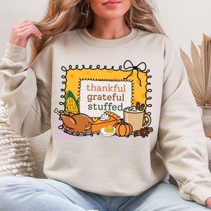May include: Beige sweatshirt with a Thanksgiving-themed graphic. The design features a yellow frame with the words "thankful, grateful, stuffed" and illustrations of a turkey, pie, corn, pumpkin, and coffee.
