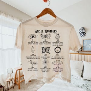 以下が含まれることがあります： 「Angel Numbers」の文字と、さまざまなシンボルと数字が黒でプリントされたクリーム色のTシャツ。目、蝶、クリスタル、花などのイラストと、インスピレーションを与えるテキストが特徴です。木製のハンガーにかかっています。