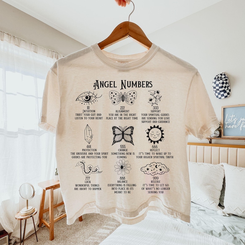以下が含まれることがあります： 「Angel Numbers」の文字と、さまざまなシンボルと数字が黒でプリントされたクリーム色のTシャツ。目、蝶、クリスタル、花などのイラストと、インスピレーションを与えるテキストが特徴です。木製のハンガーにかかっています。