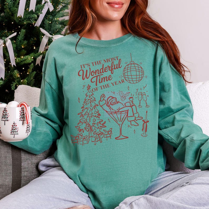 Puede incluir: Una sudadera verde azulado con la frase "It's the Most Wonderful Time of the Year" en letras rojas. El gr&aacute;fico presenta un &aacute;rbol de Navidad, Pap&aacute; Noel en una copa de martini y una bola de discoteca.