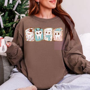 May include: Brown long-sleeve sweatshirt featuring a graphic of four cartoon teeth, each with a different holiday-themed design. One tooth wears a Santa hat, another has reindeer antlers, and the others have a scarf and a winter hat. The sweatshirt is a solid brown color.