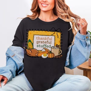 May include: Black sweatshirt with a Thanksgiving-themed graphic. The design features the words "thankful, grateful, stuffed" inside a yellow frame, along with illustrations of a turkey, pie, pumpkin, and coffee.