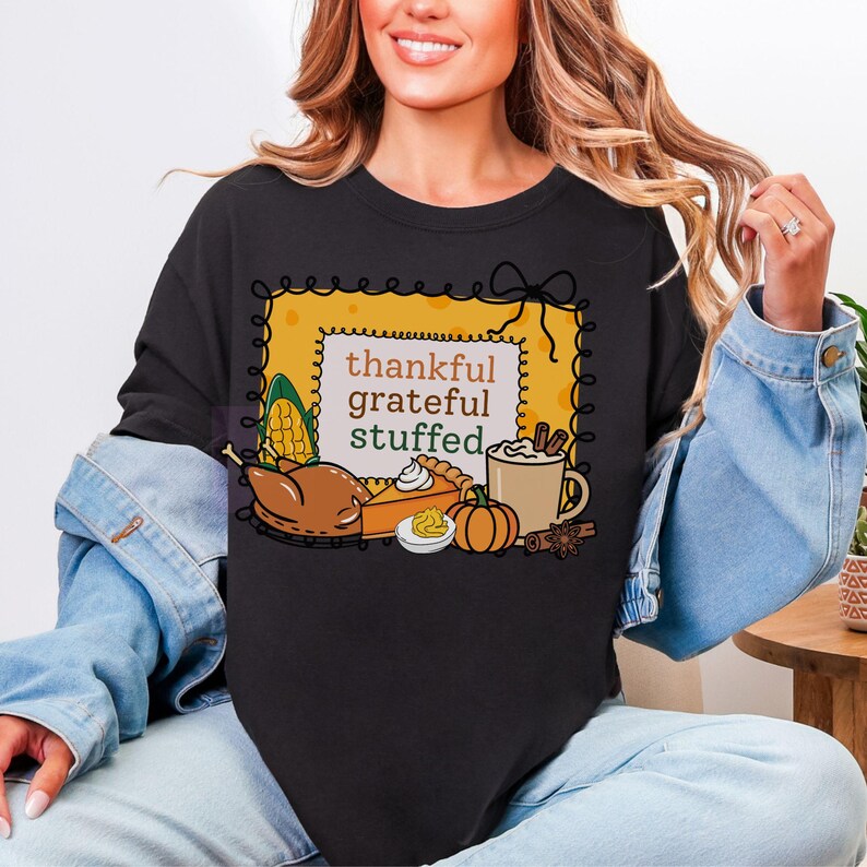 May include: Black sweatshirt with a Thanksgiving-themed graphic. The design features the words "thankful, grateful, stuffed" inside a yellow frame, along with illustrations of a turkey, pie, pumpkin, and coffee.