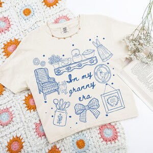 May include: Cream-colored t-shirt with a blue graphic design featuring a shelf with teapots, a cat, a bow, and the text "In my granny era." The shirt is laid on a colorful crocheted blanket.