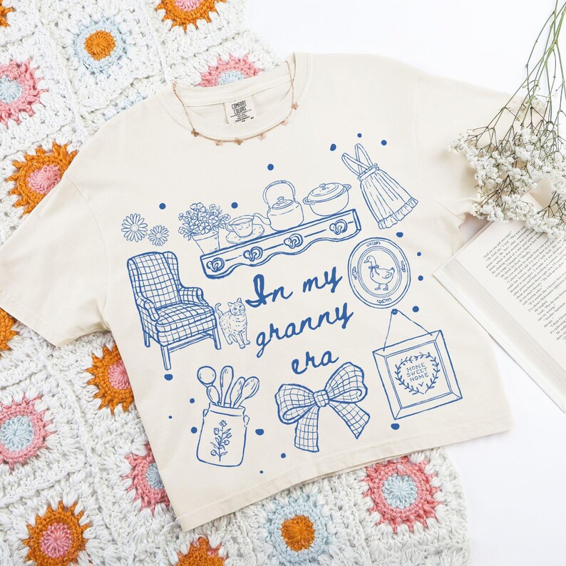 May include: Cream-colored t-shirt with a blue graphic design featuring a shelf with teapots, a cat, a bow, and the text "In my granny era." The shirt is laid on a colorful crocheted blanket.