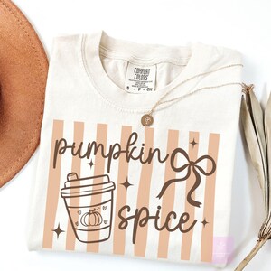 May include: Cream-colored t-shirt with a "pumpkin spice" graphic. The design includes a coffee cup with a pumpkin, a bow, and vertical peach stripes. A gold necklace with a pendant is draped on the shirt.