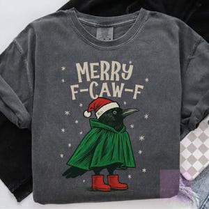 Może przedstawiać: Ciemnoszara bluza z napisem "MERRY F-CAW-F" i rysunkiem kruka w czapce Świętego Mikołaja, zielonej pelerynie i czerwonych butach. Białe płatki śniegu otaczają kruka. Bluza ma luźny krój.
