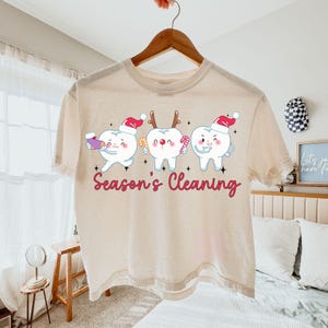 Puede incluir: Una camiseta beige con un tema dental festivo. La camiseta presenta tres dientes de dibujos animados con gorros de Pap&aacute; Noel y sosteniendo art&iacute;culos navide&ntilde;os. El texto "Season's Cleaning" est&aacute; impreso en cursiva debajo. La camiseta est&aacute; colgada en una percha de madera.