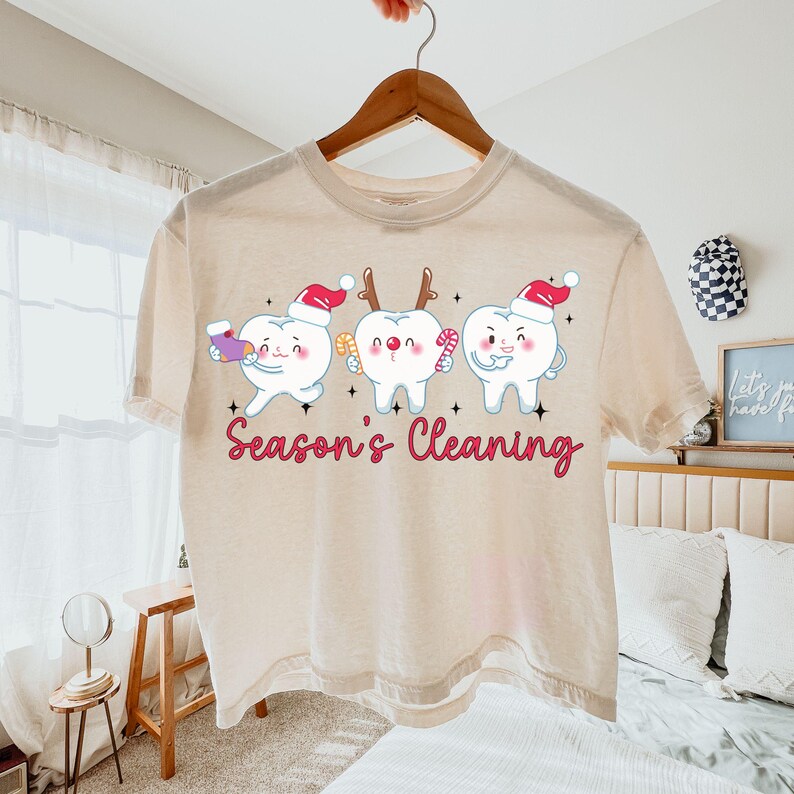 Puede incluir: Una camiseta beige con un tema dental festivo. La camiseta presenta tres dientes de dibujos animados con gorros de Pap&aacute; Noel y sosteniendo art&iacute;culos navide&ntilde;os. El texto "Season's Cleaning" est&aacute; impreso en cursiva debajo. La camiseta est&aacute; colgada en una percha de madera.