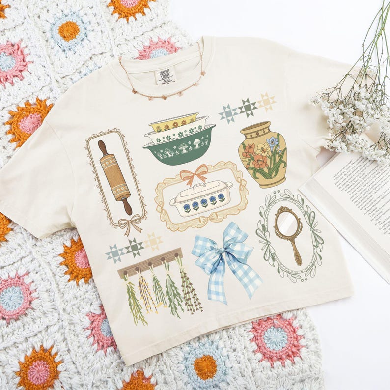 May include: Cream-colored t-shirt featuring vintage kitchen-themed illustrations, including a rolling pin, bowls, a vase, and a blue gingham bow. The shirt is adorned with decorative frames and starburst accents, evoking a retro aesthetic.