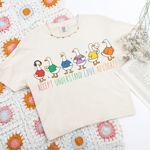 May include: Cream-colored t-shirt featuring a line of cartoon geese, each wearing different colored outfits. The text "ACCEPT UNDERSTAND LOVE ADVOCATE" is printed below the geese in rainbow colors. A crochet blanket and a book are in the background.