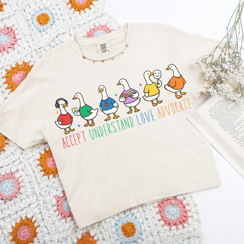 May include: Cream-colored t-shirt featuring a line of cartoon geese, each wearing different colored outfits. The text "ACCEPT UNDERSTAND LOVE ADVOCATE" is printed below the geese in rainbow colors. A crochet blanket and a book are in the background.