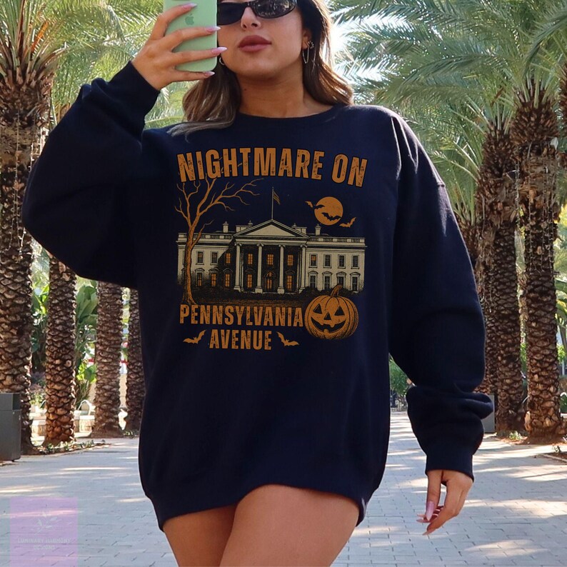 K&ouml;nnte beinhalten: Marineblaues Sweatshirt mit einem Halloween-Motiv. Das Design zeigt den Text "NIGHTMARE ON PENNSYLVANIA AVENUE" &uuml;ber einer Illustration des Wei&szlig;en Hauses, eines K&uuml;rbisses, eines Baumes, eines Mondes und Flederm&auml;usen.