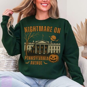 K&ouml;nnte beinhalten: Dunkelgr&uuml;nes Sweatshirt mit einem Halloween-Motiv. Das Design zeigt den Text "NIGHTMARE ON PENNSYLVANIA AVENUE" &uuml;ber einer Illustration des Wei&szlig;en Hauses, eines K&uuml;rbisses, eines Baumes, eines Mondes und Flederm&auml;usen.