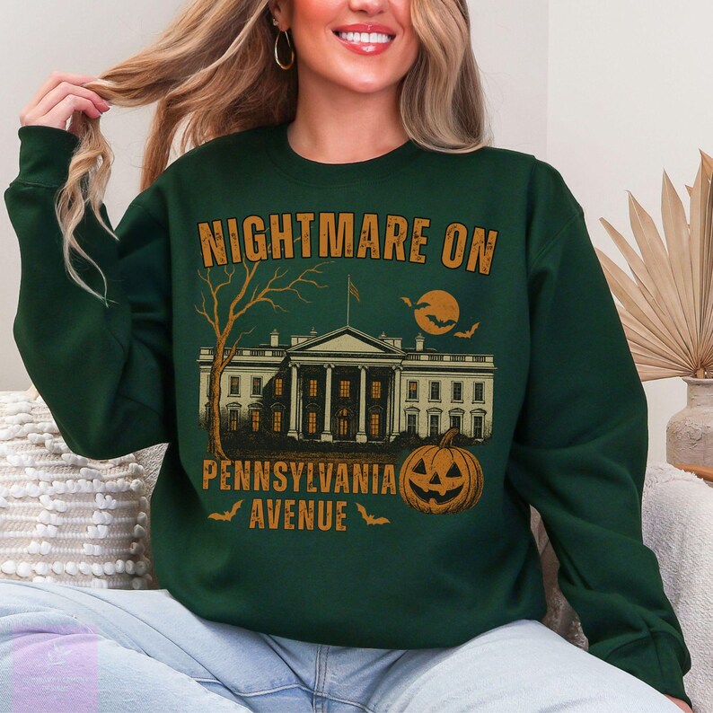 K&ouml;nnte beinhalten: Dunkelgr&uuml;nes Sweatshirt mit einem Halloween-Motiv. Das Design zeigt den Text "NIGHTMARE ON PENNSYLVANIA AVENUE" &uuml;ber einer Illustration des Wei&szlig;en Hauses, eines K&uuml;rbisses, eines Baumes, eines Mondes und Flederm&auml;usen.