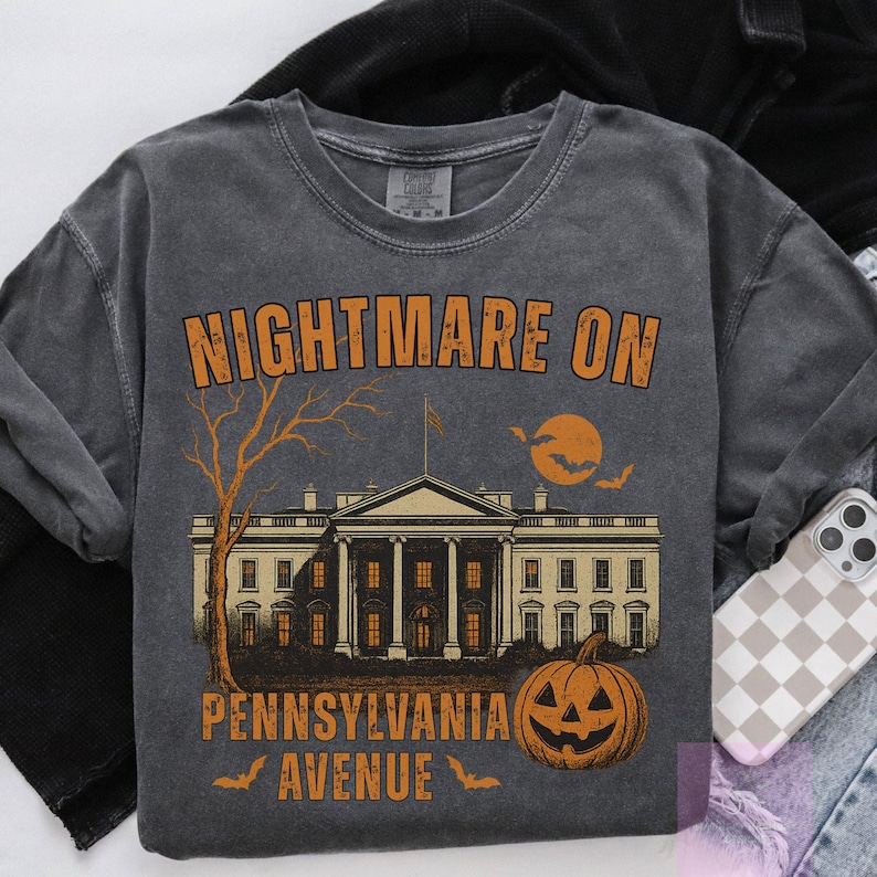 K&ouml;nnte beinhalten: Dunkelgraues Sweatshirt mit dem Schriftzug "NIGHTMARE ON PENNSYLVANIA AVENUE" in Orange. Das Design zeigt eine gruselige Illustration des Wei&szlig;en Hauses, einen kahlen Baum, einen K&uuml;rbis, einen Vollmond und Flederm&auml;use, ideal f&uuml;r Halloween.