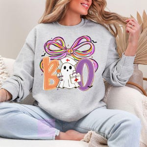 Pu&ograve; includere: Felpa grigio chiaro con grafica a tema Halloween. Il design include la parola "BOO" con un fantasma che indossa un cappello da infermiera e tiene una siringa e una borsa medica. Un fiocco colorato e pipistrelli completano il design.