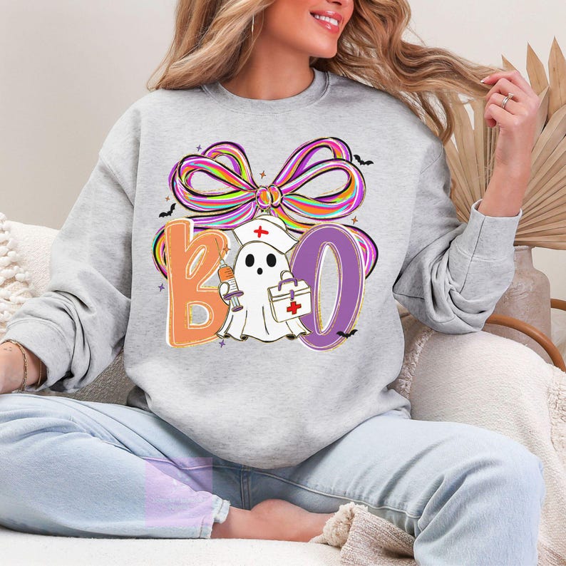 Pu&ograve; includere: Felpa grigio chiaro con grafica a tema Halloween. Il design include la parola "BOO" con un fantasma che indossa un cappello da infermiera e tiene una siringa e una borsa medica. Un fiocco colorato e pipistrelli completano il design.
