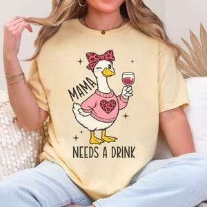 Peut inclure: T-shirt jaune clair avec un dessin d'oie de dessin anim&eacute; portant un pull rose et un n&oelig;ud, tenant un verre de vin. Le texte "MAMA NEEDS A DRINK" est imprim&eacute; en dessous. Le design est en noir et rose.