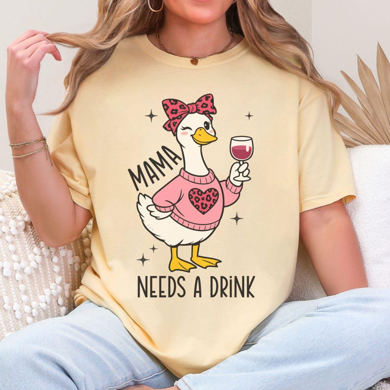 Peut inclure: T-shirt jaune clair avec un dessin d'oie de dessin anim&eacute; portant un pull rose et un n&oelig;ud, tenant un verre de vin. Le texte "MAMA NEEDS A DRINK" est imprim&eacute; en dessous. Le design est en noir et rose.