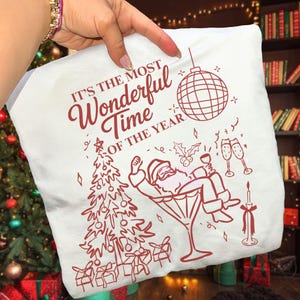 Puede incluir: Funda de almohada blanca con un gr&aacute;fico rojo festivo. El texto dice "IT'S THE MOST Wonderful Time OF THE YEAR." El dise&ntilde;o incluye una bola de discoteca, un &aacute;rbol de Navidad, Pap&aacute; Noel en una copa de Martini, copas de champ&aacute;n y una vela.