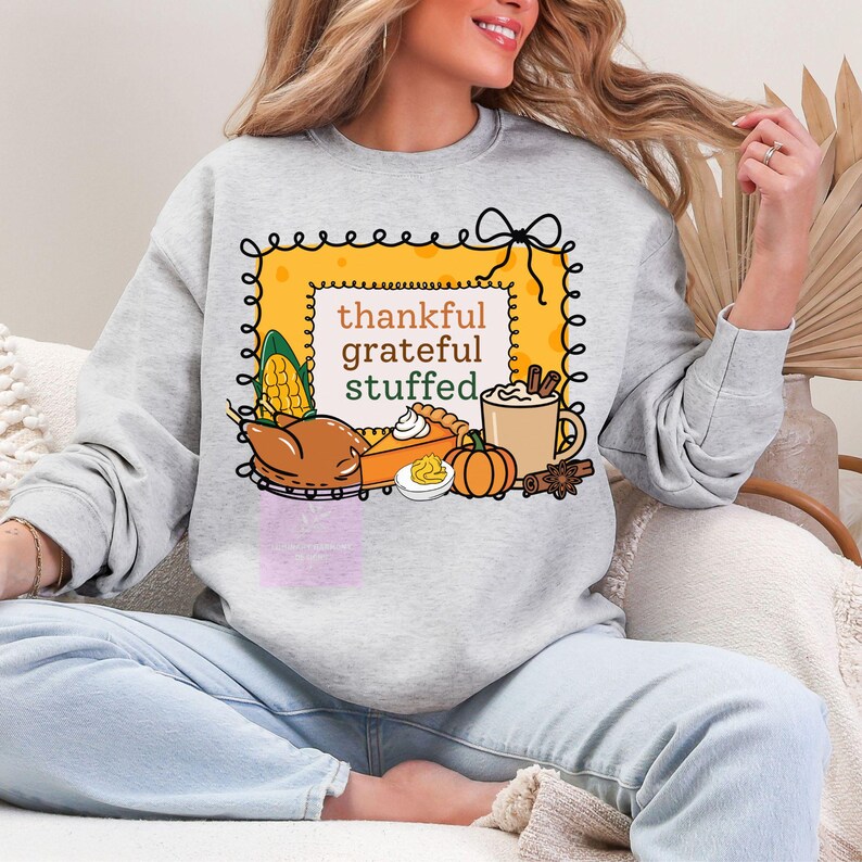 May include: A light gray sweatshirt featuring a Thanksgiving-themed graphic. The graphic includes the words "thankful, grateful, stuffed" inside a yellow frame, along with illustrations of a turkey, pie, pumpkin, and corn.