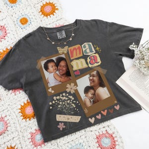 May include: Dark grey t-shirt with the word "mama" in colourful letters and two framed photos. The shirt features a floral design and a gold necklace. The shirt is displayed on a crocheted blanket.