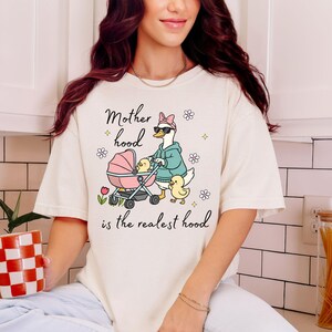 May include: Cream-colored t-shirt with the phrase "Motherhood is the realest hood." The graphic features a goose pushing a pink stroller with ducklings. The goose wears sunglasses and a hoodie. Floral and diamond accents surround the text and graphic.