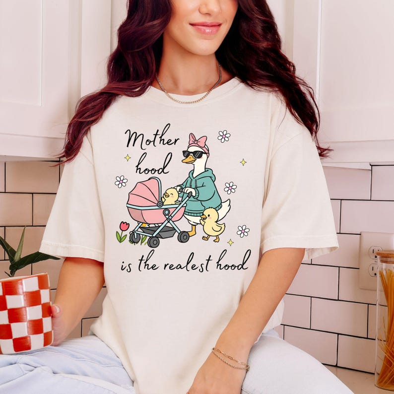 May include: Cream-colored t-shirt with the phrase "Motherhood is the realest hood." The graphic features a goose pushing a pink stroller with ducklings. The goose wears sunglasses and a hoodie. Floral and diamond accents surround the text and graphic.