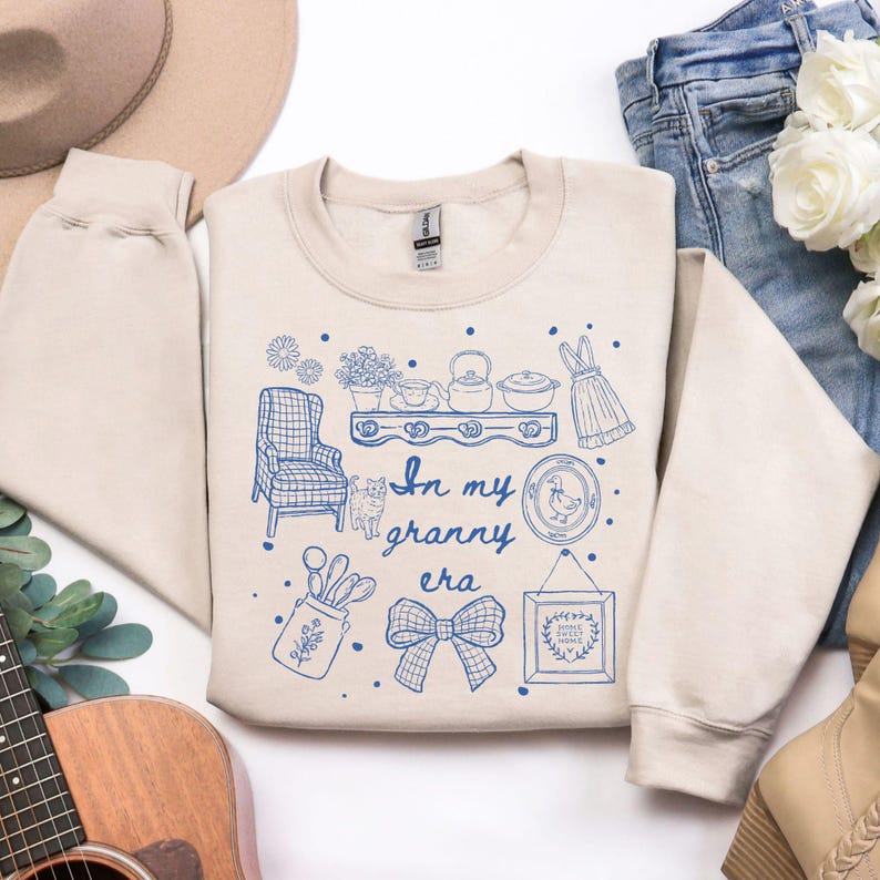 May include: Beige sweatshirt with a blue graphic design featuring various vintage-style illustrations, including a chair, cat, and kitchenware. The text "In my granny era" is also included in the design. The sweatshirt is folded.