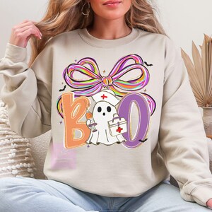 Pu&ograve; includere: Felpa beige con un fiocco colorato e la scritta "BOO" in arancione e viola. Un fantasma con un cappello da infermiera e con una siringa e una borsa medica &egrave; al centro. Piccoli pipistrelli neri sono sparsi intorno al disegno.
