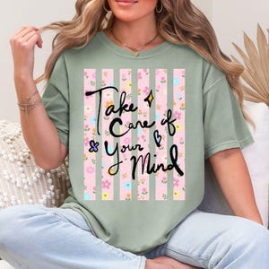 May include: Sage green t-shirt with the text "Take Care of Your Mind" in black script. The shirt features a pink and white striped design with floral accents. The shirt is a relaxed fit.