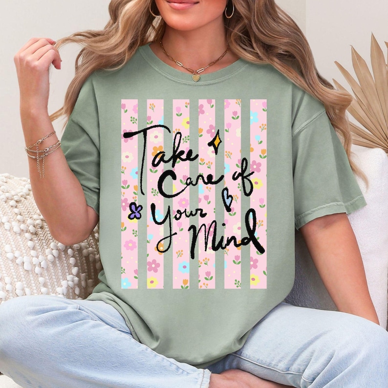 May include: Sage green t-shirt with the text "Take Care of Your Mind" in black script. The shirt features a pink and white striped design with floral accents. The shirt is a relaxed fit.